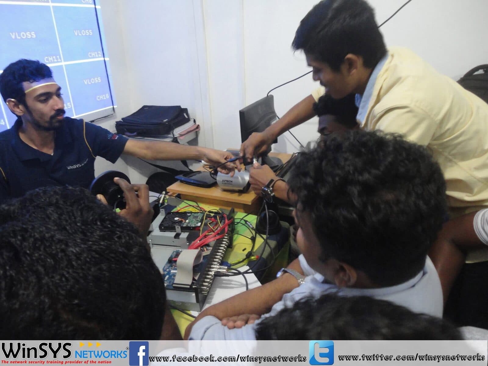An instructor demonstrating CCTV camera installation to students around a workbench